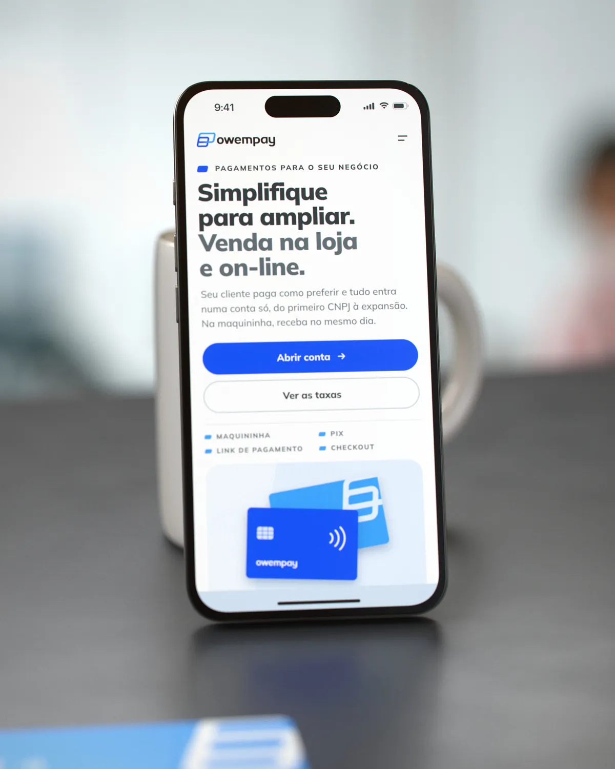 Celular em pé, encostado numa caneca de cerâmica sobre o balcão, com a primeira dobra da landing page da OwemPay; ao fundo, desfocado, o balcão de um café
