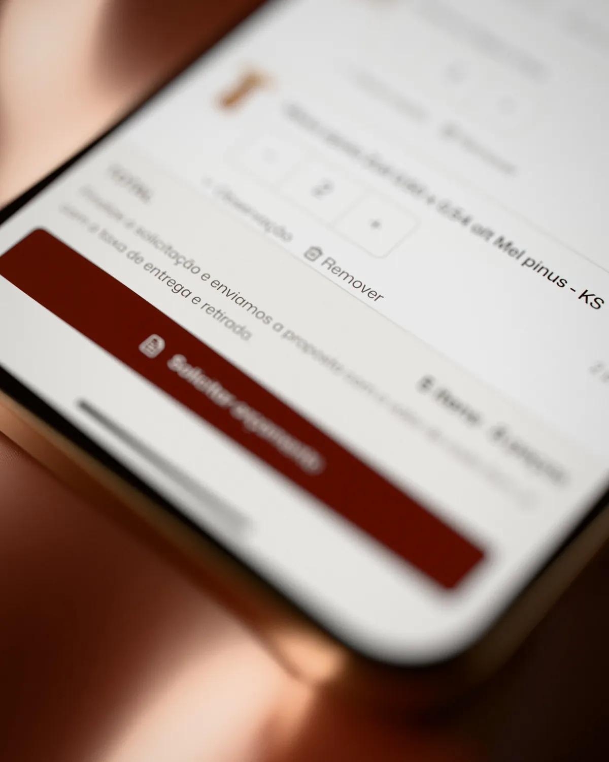 Close de um celular sobre cetim vinho: a relação de itens com o contador de quantidade e o botão Solicitar orçamento